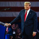 Trump’s Michigan Power Play Puts Democrats on Defense Man in suit at podium with American flag backdrop
