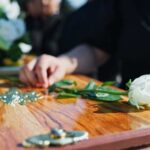 A hand placing a white rose on a wooden casket during a funeral service