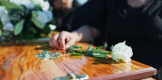 A hand placing a white rose on a wooden casket during a funeral service